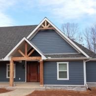 Gray house exterior with dark roof, front porch, and exposed wood trusses under a partly cloudy sky
