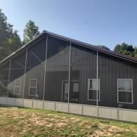 Dark gray building with a metal roof, large windows, and a concrete foundation under a clear blue sky.
