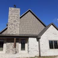 Suburban house with stone chimney, gray siding, and a steep gabled roof under a blue sky