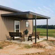 Small metal porch with bench and chairs beside a tan building in a rural field.