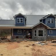 Partially built blue house with stone facade and muddy construction site under cloudy sky