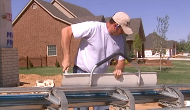 Worker in white shirt and cap measuring metal roofing panels outdoors beside brick houses