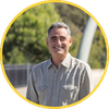 Man smiling outdoors, wearing a striped button-down shirt, framed in a yellow circular border.