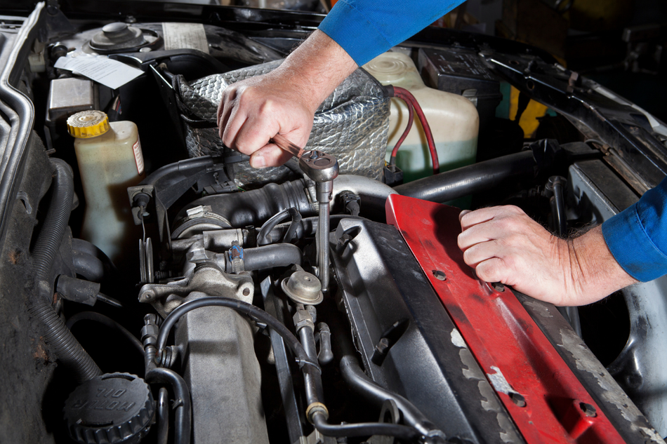 Hands working on a car engine in a garage, with a red engine cover and tools nearby