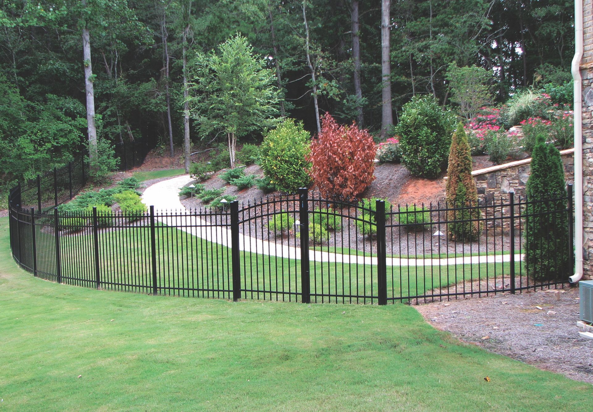 Black metal fence curves around a green lawn leading to a landscaped garden path.