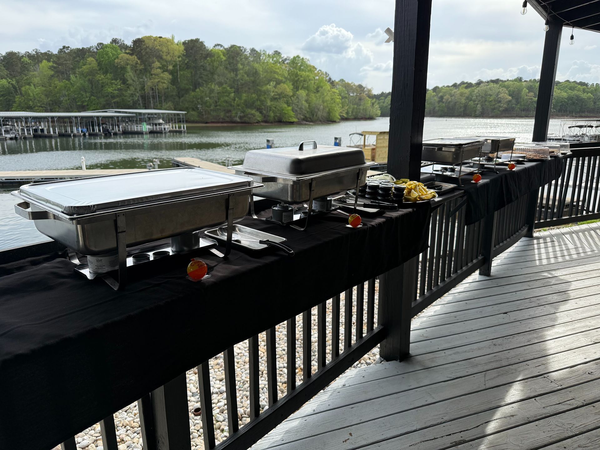 Outdoor buffet setup on a deck overlooking a lake. Food warmers and serving utensils are arranged on black tablecloths.