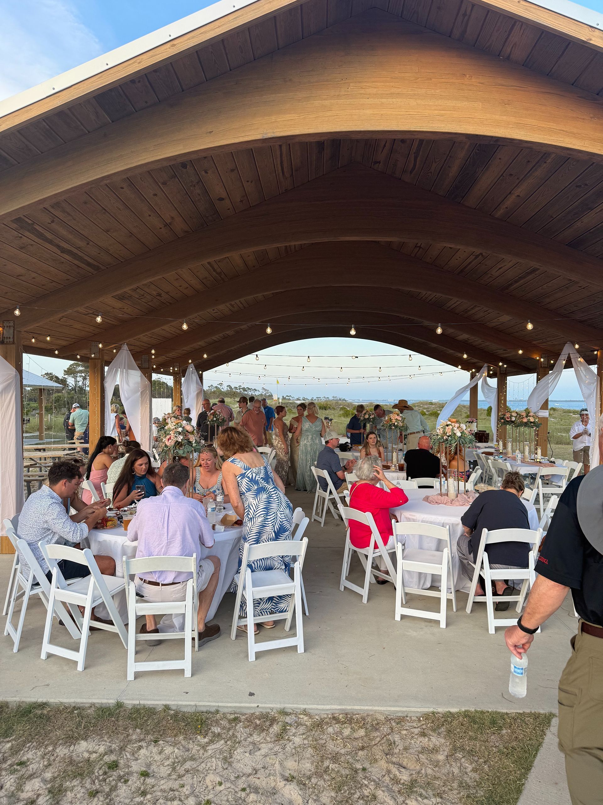 Outdoor event under a wooden pavilion. People seated at tables, eating and socializing. White chairs, string lights.