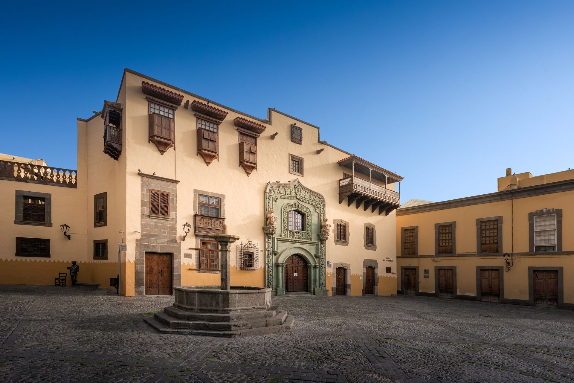 Historic plaza with a ornate stone fountain in front of beige colonial buildings under a blue sky, Gran Canaria