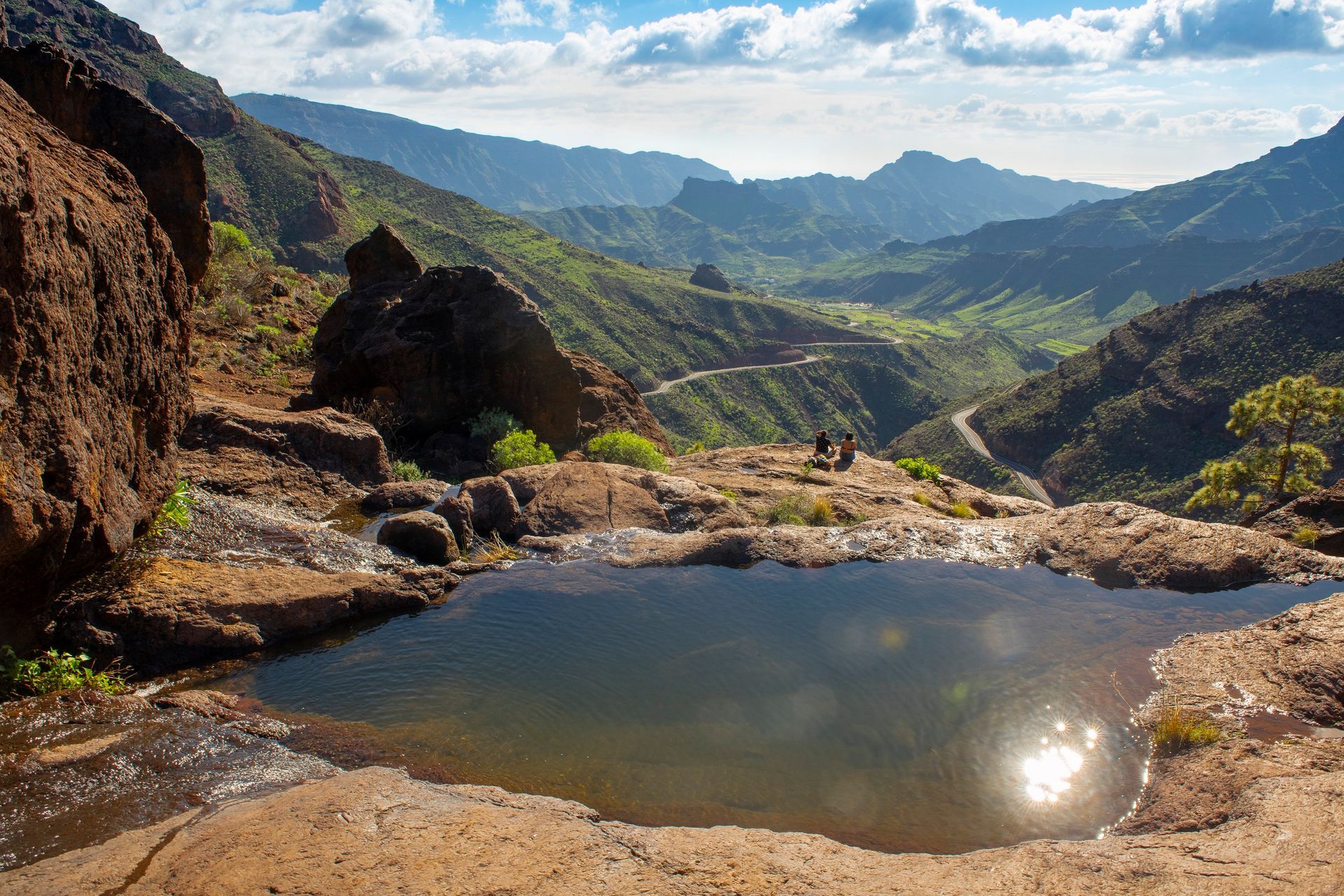 Rocky mountain overlook with a small reflective pool and sunlit green valley under a cloudy sky. Los Azulejos, Gran Canaria
