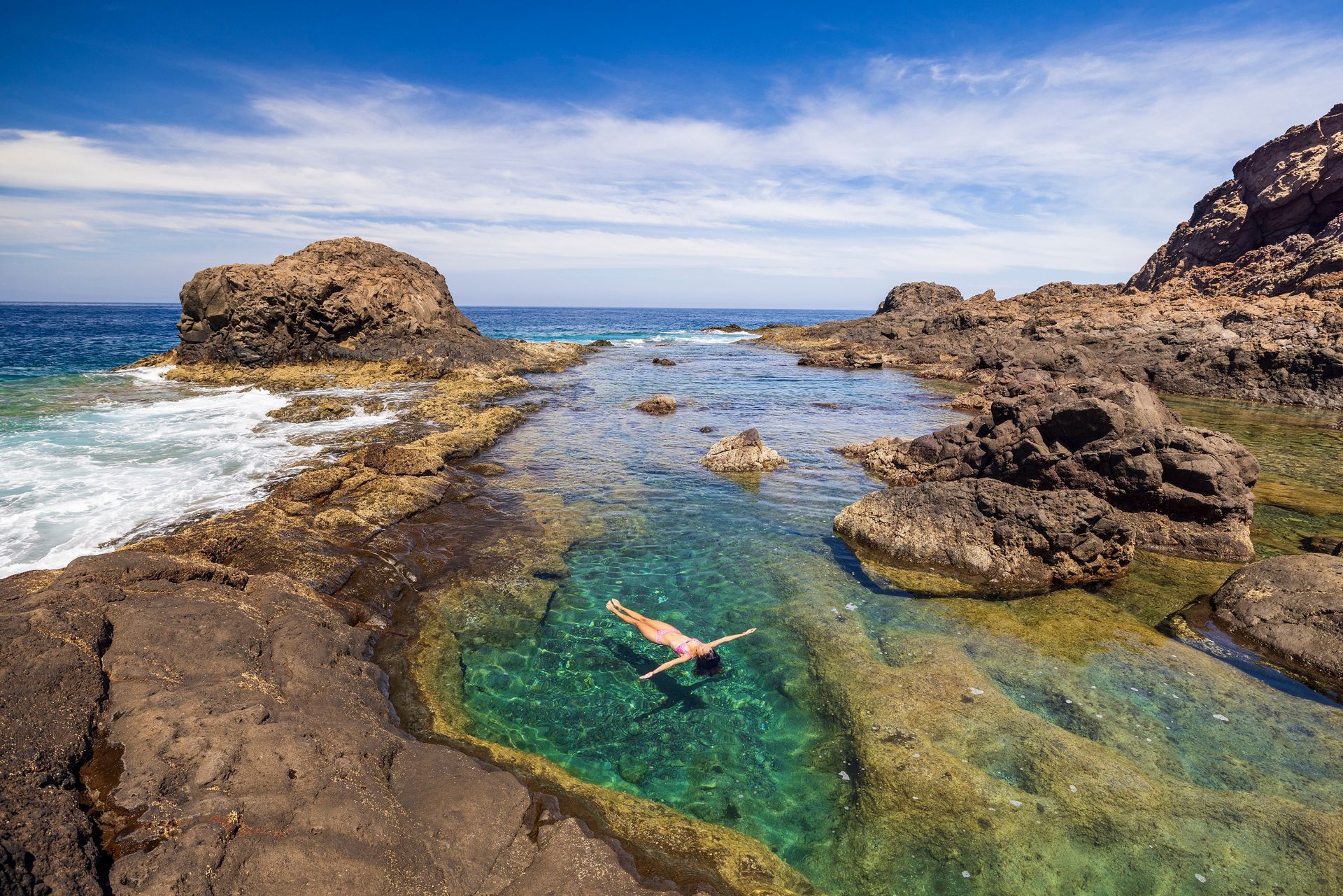 Person floating in a clear green tide pool among rocky coastal cliffs under a blue sky. Natural pools, Gran Canaria.