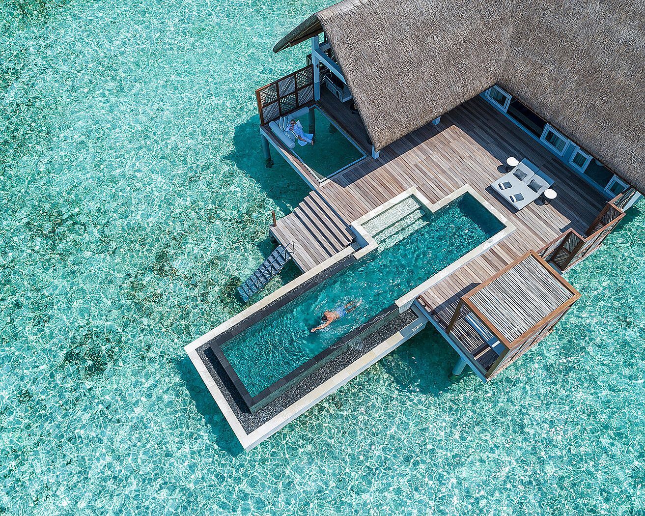 Overwater bungalow with thatched roof, pool, and a person swimming in turquoise water.