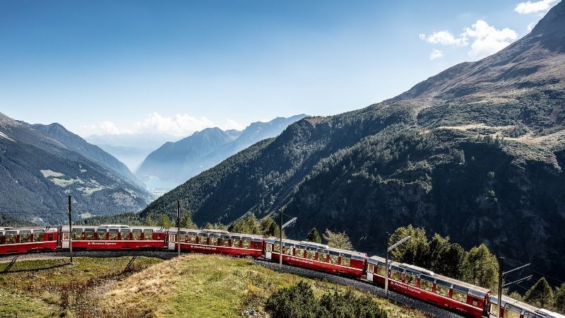 Red train curving through a mountain valley with a lake and snow-capped peaks in the background