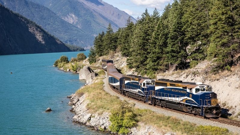 Blue passenger train curves along a lakeside track beside turquoise water and pine-covered mountains.