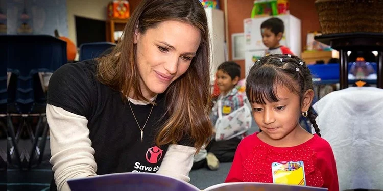 Woman reading a book with a young girl. Several other children are in the room.