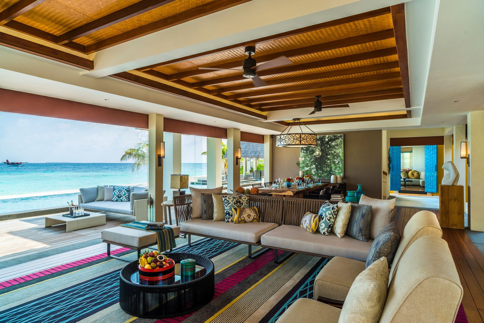 Spacious beachfront living room with ocean view, colourful rug, neutral-toned furniture, and wooden ceiling.