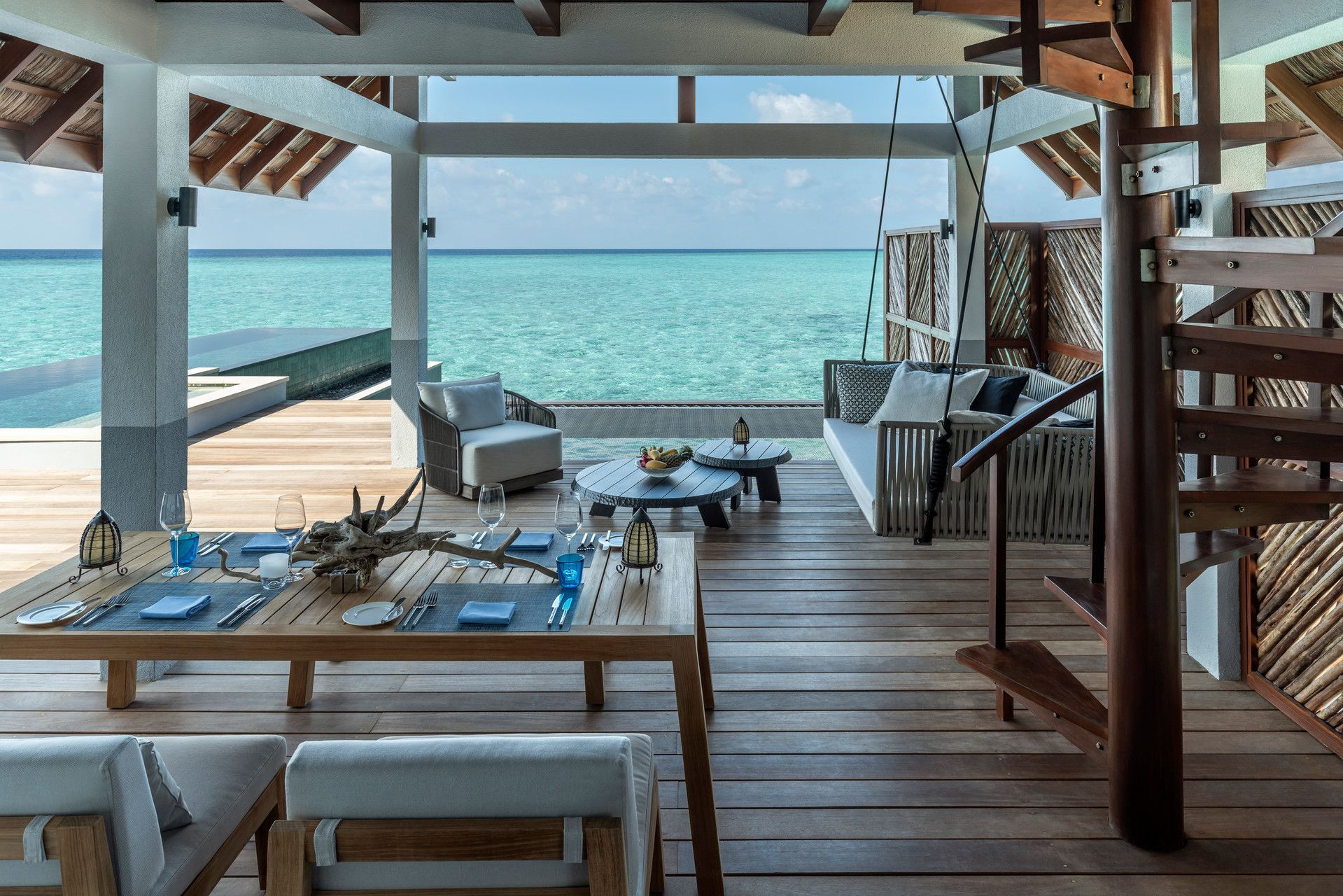 Outdoor deck with dining and lounge area, overlooking turquoise ocean. Wooden staircase and carved screen.