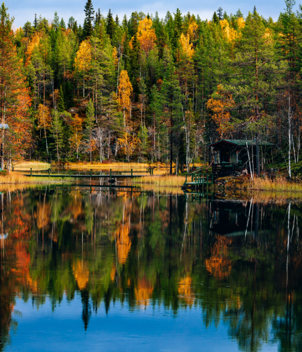 Autumn forest reflected in a calm lake, with green, yellow, and orange trees and a small dark cabin