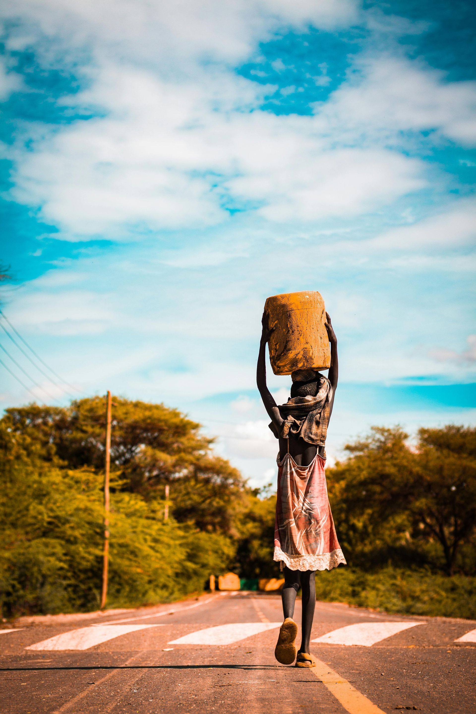 Person walking down a road, carrying a large object on their head. Bright blue sky, trees on both sides.