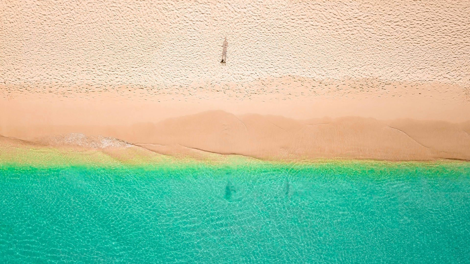 Aerial view of a beach with turquoise water, a sandy shore, and one person standing at the edge.