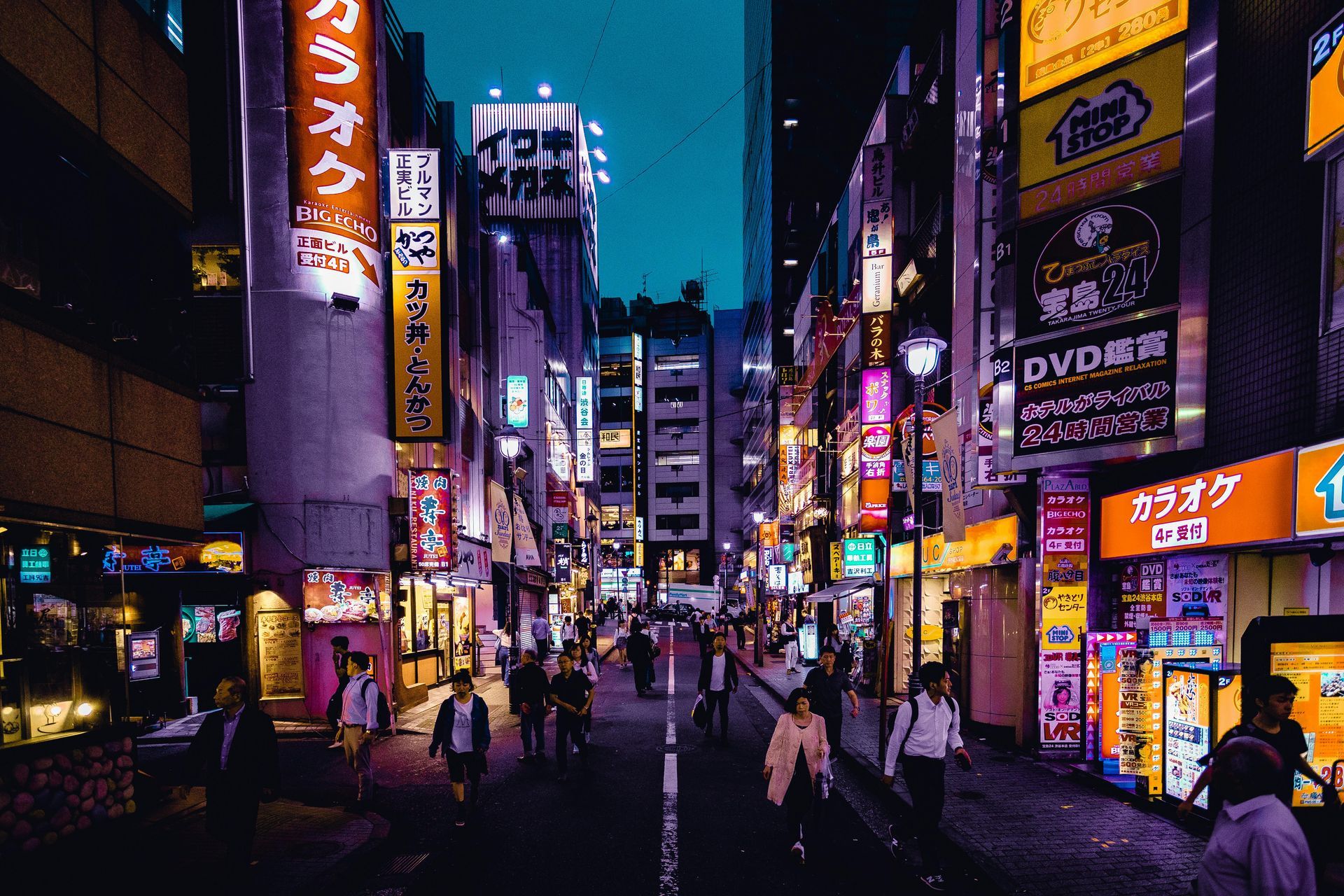 Busy Tokyo street at dusk, neon signs illuminate buildings. People walk along the street.