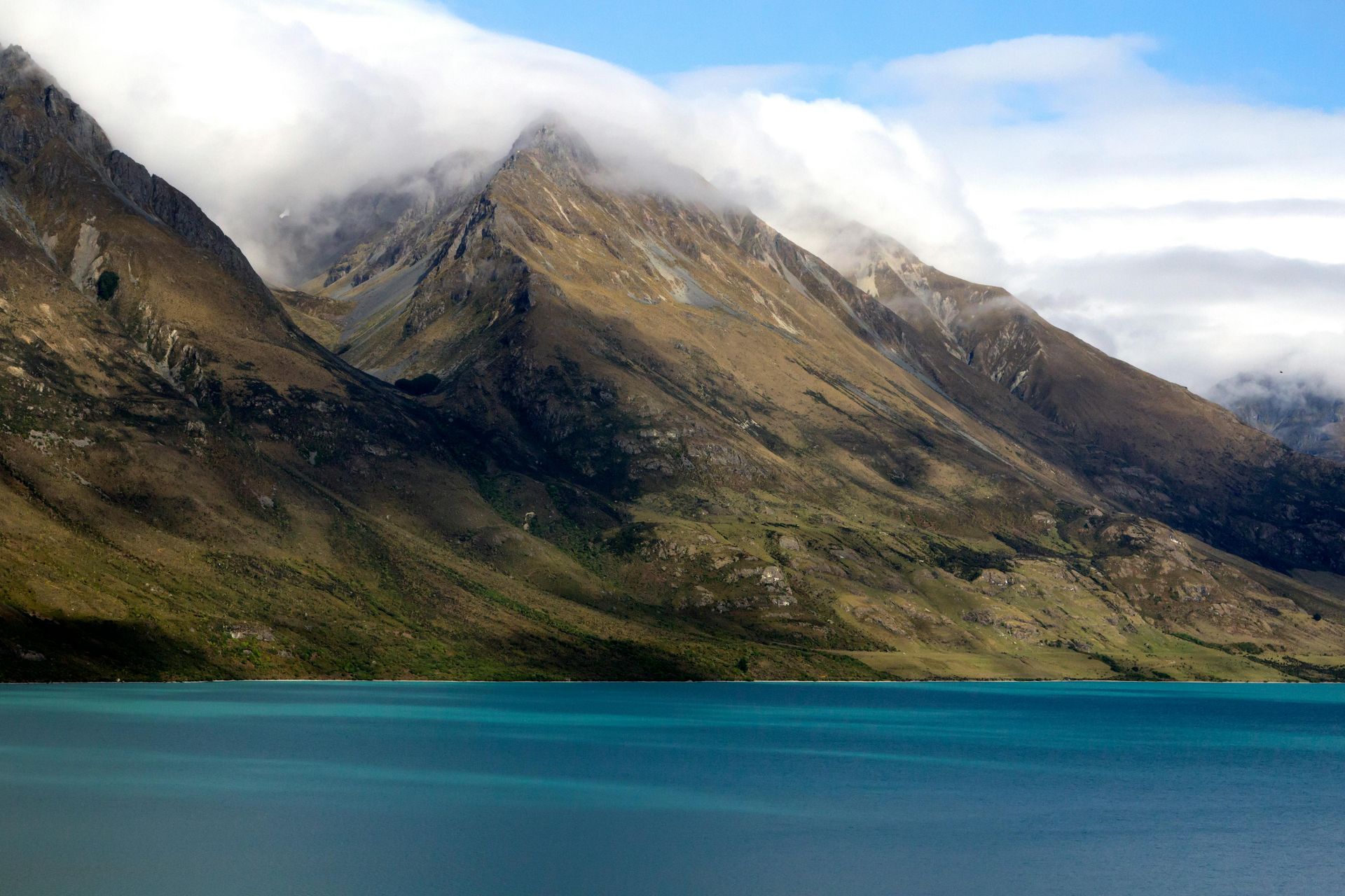 Turquoise lake with mountains partially obscured by clouds. Brown and green landscape.