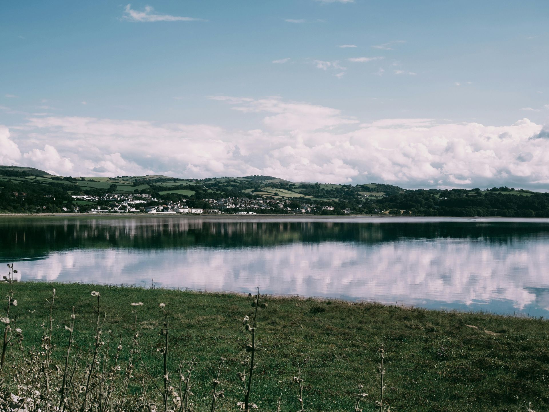Cornwall. Calm lake reflecting white clouds and a distant town under a blue sky. Green grass in the foreground.