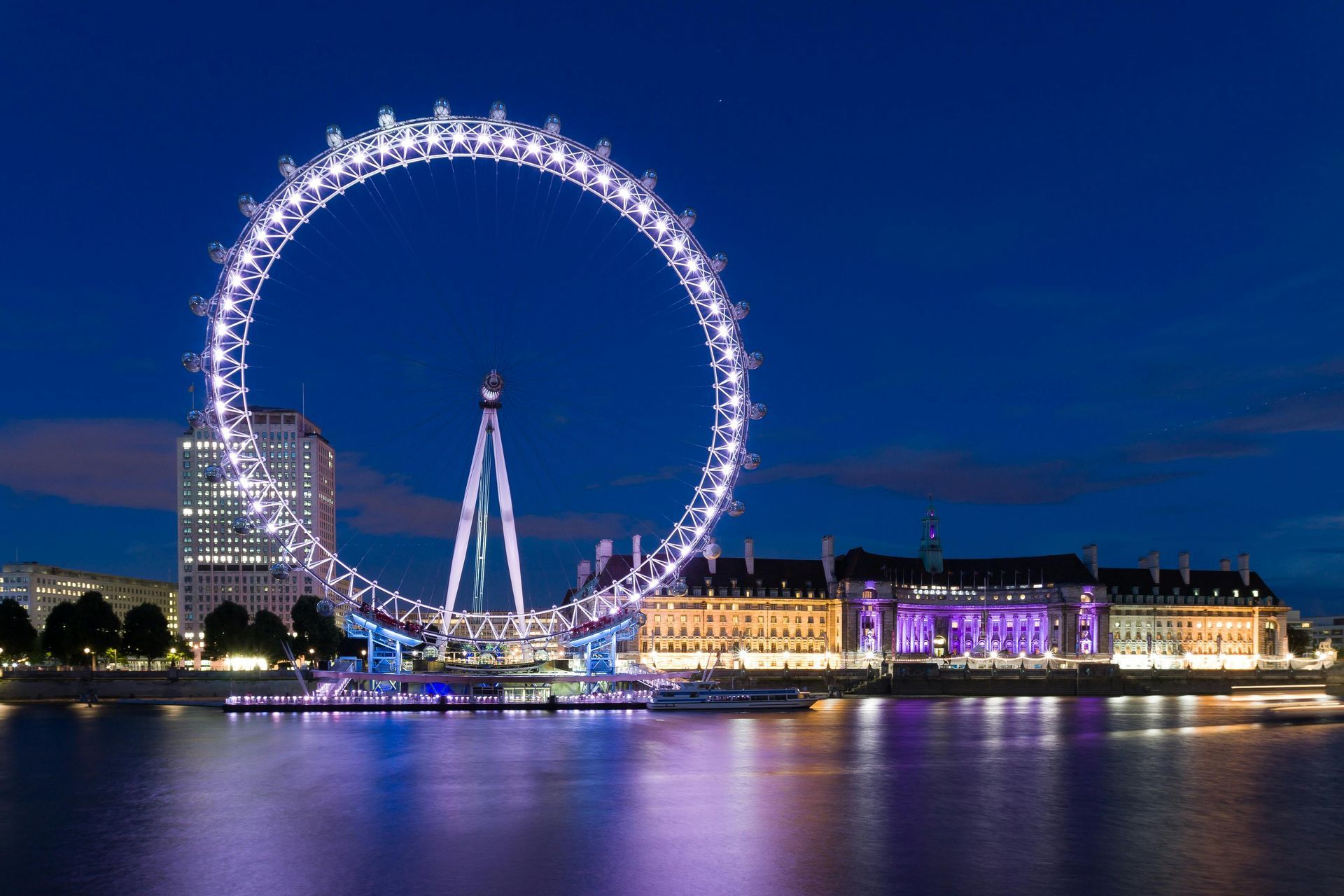 London Eye Ferris wheel lit up at night, reflecting in the Thames River.