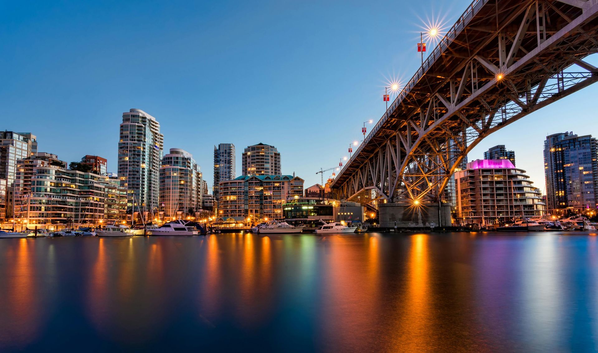 City skyline at dusk with lit buildings, bridge, and harbor. Water reflects lights.