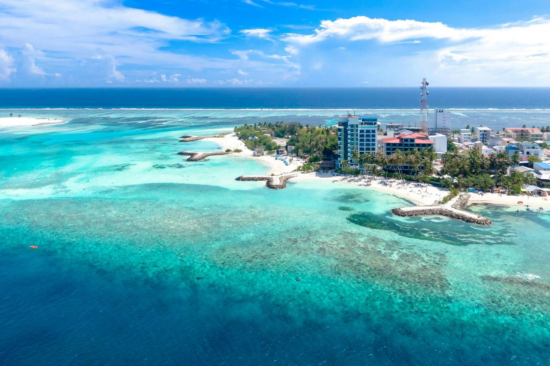 Aerial view of turquoise ocean surrounding a small island with buildings and a beach under a blue sky.