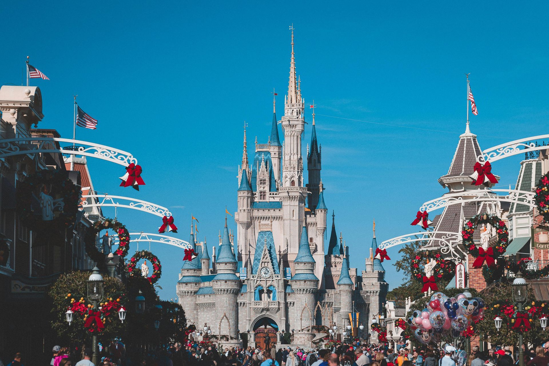Cinderella Castle at Magic Kingdom, decorated for Christmas. Crowd of people on Main Street, USA, under a bright blue sky.