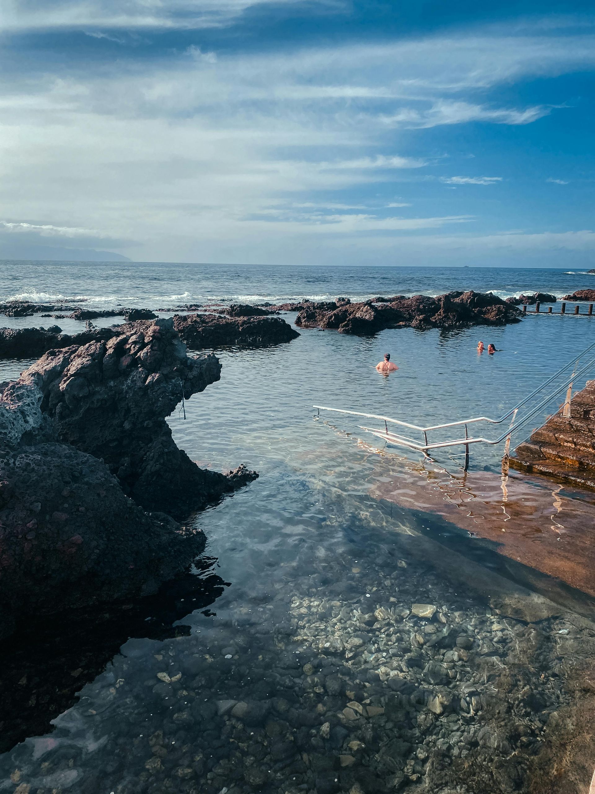Rocky tide pools with clear water, connected to the ocean under a blue sky. People swim in the distance.