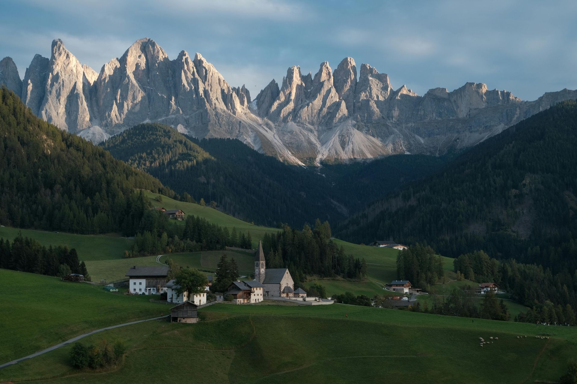 Rolling green hills, village and church with jagged mountain range backdrop at sunset.