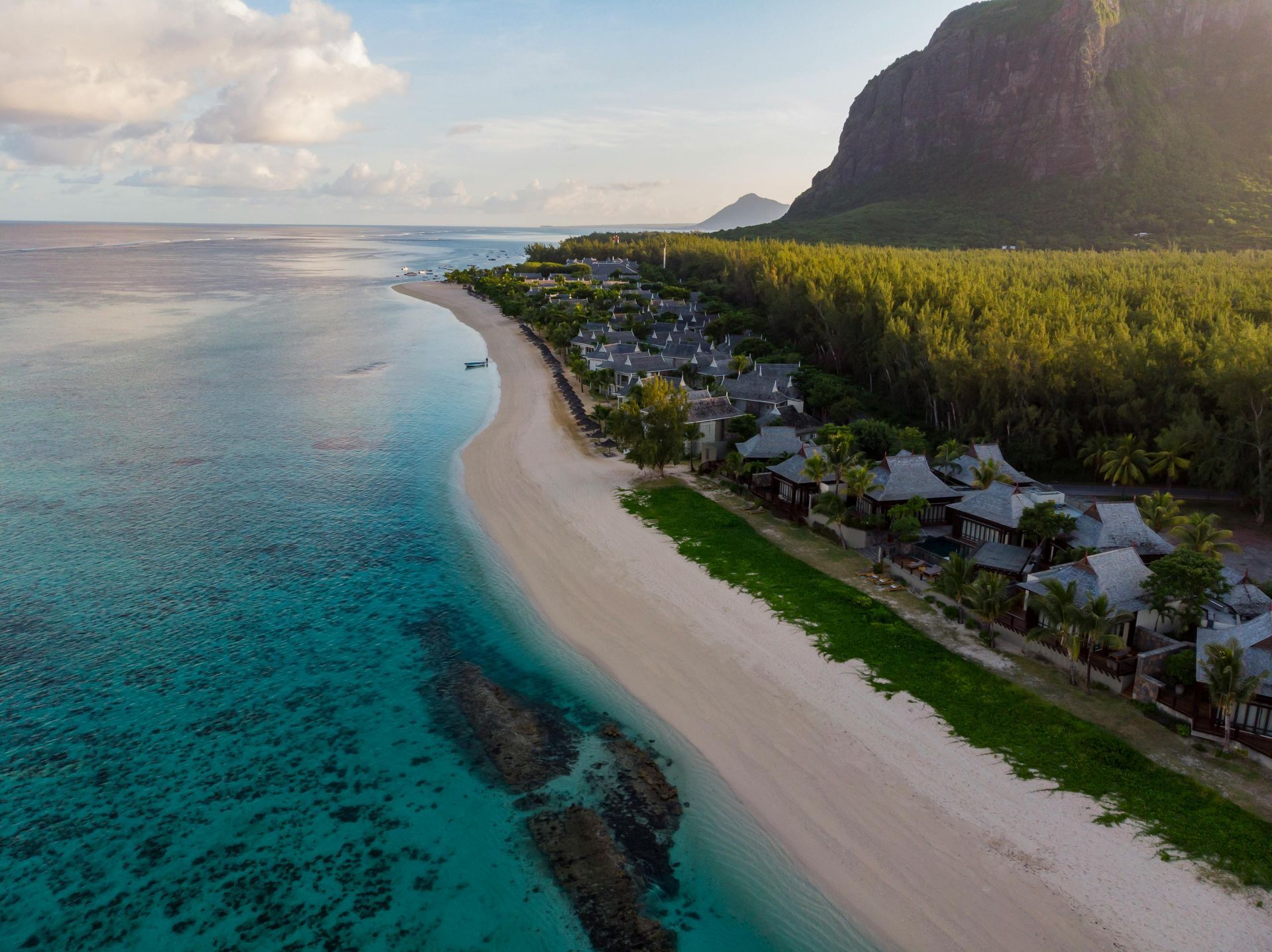 Beachside resort with white sand, turquoise water, and lush green foliage at the foot of a mountain.