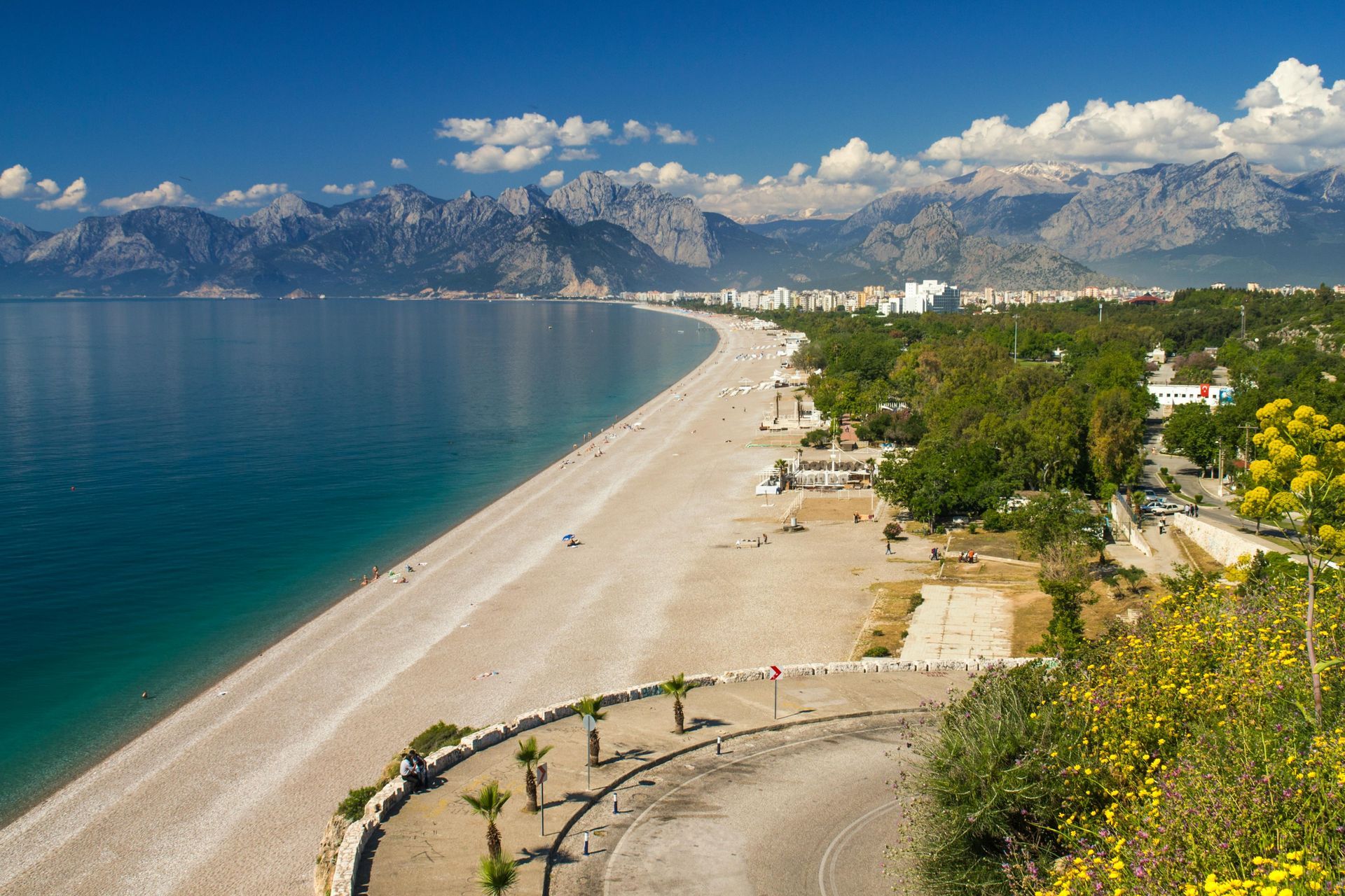 Beach scene with mountains in the background, blue water, and a coastal road.