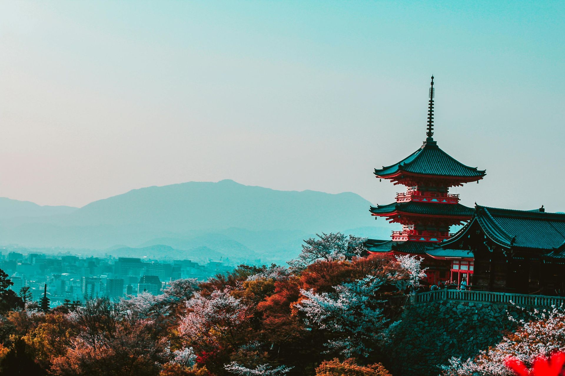Red and black Japanese pagoda on a hill, overlooking a town and mountains under a blue sky.