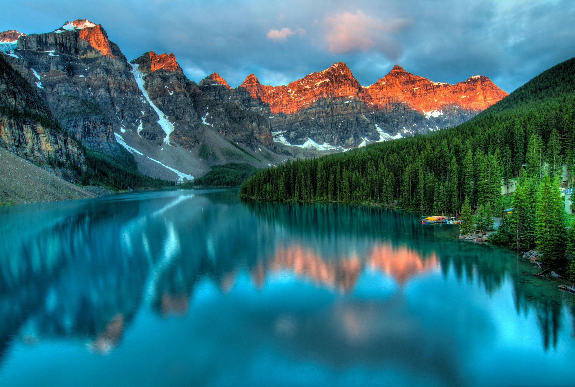 Mountains reflected in a blue lake, with a forest and colorful sky.