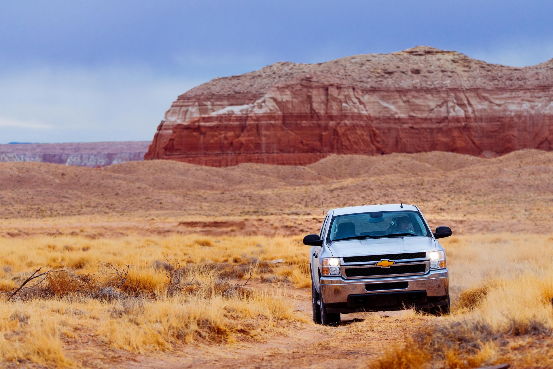 Silver pickup truck driving on a dirt road in a desert landscape, red rock butte in the background.