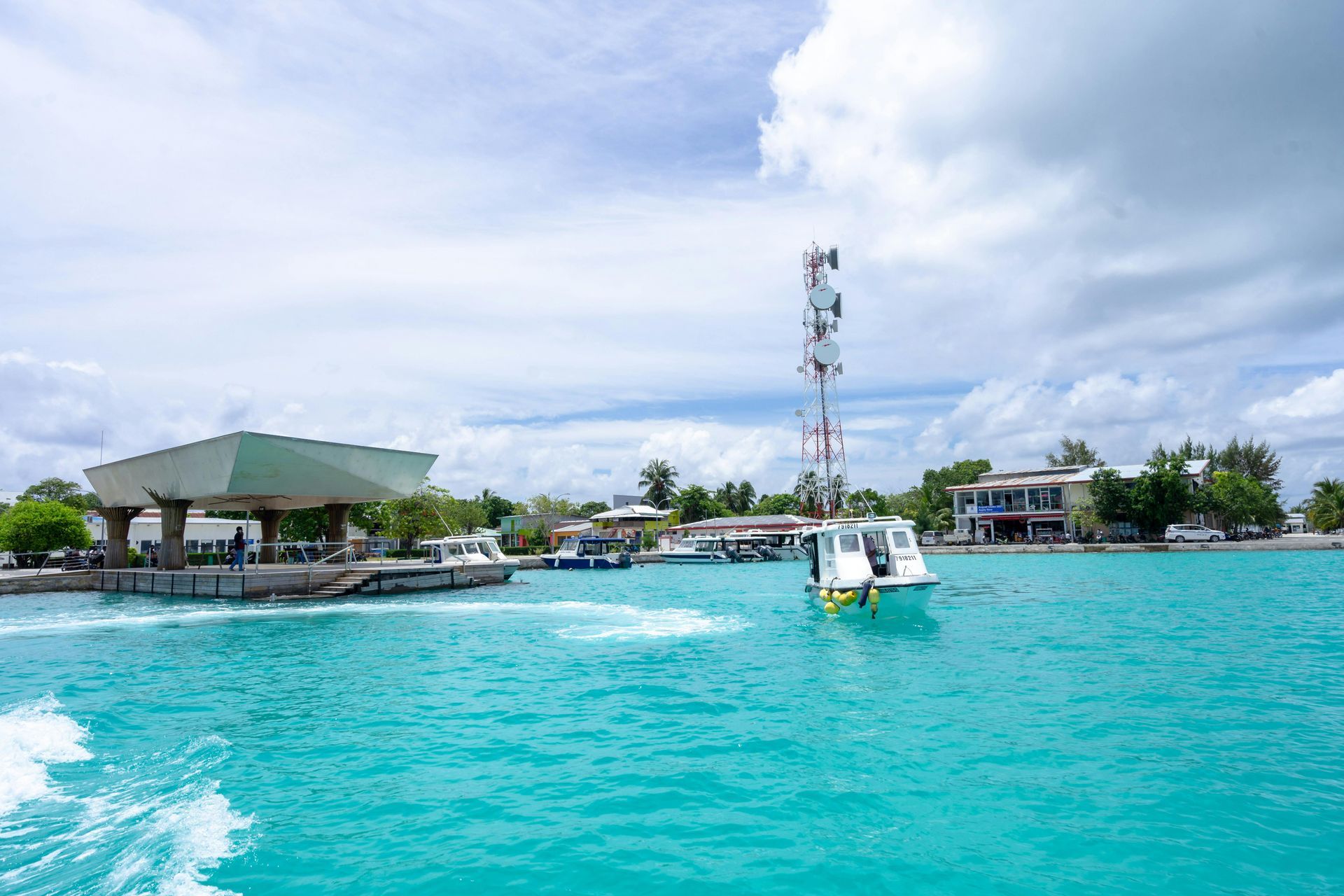 A turquoise ocean with boats, docks, buildings, and a communications tower under a cloudy sky.