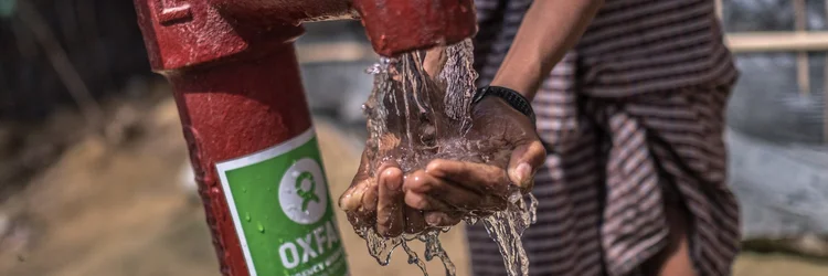 Water pours over cupped hands at a red Oxfam water pump. A person stands nearby in striped clothing.