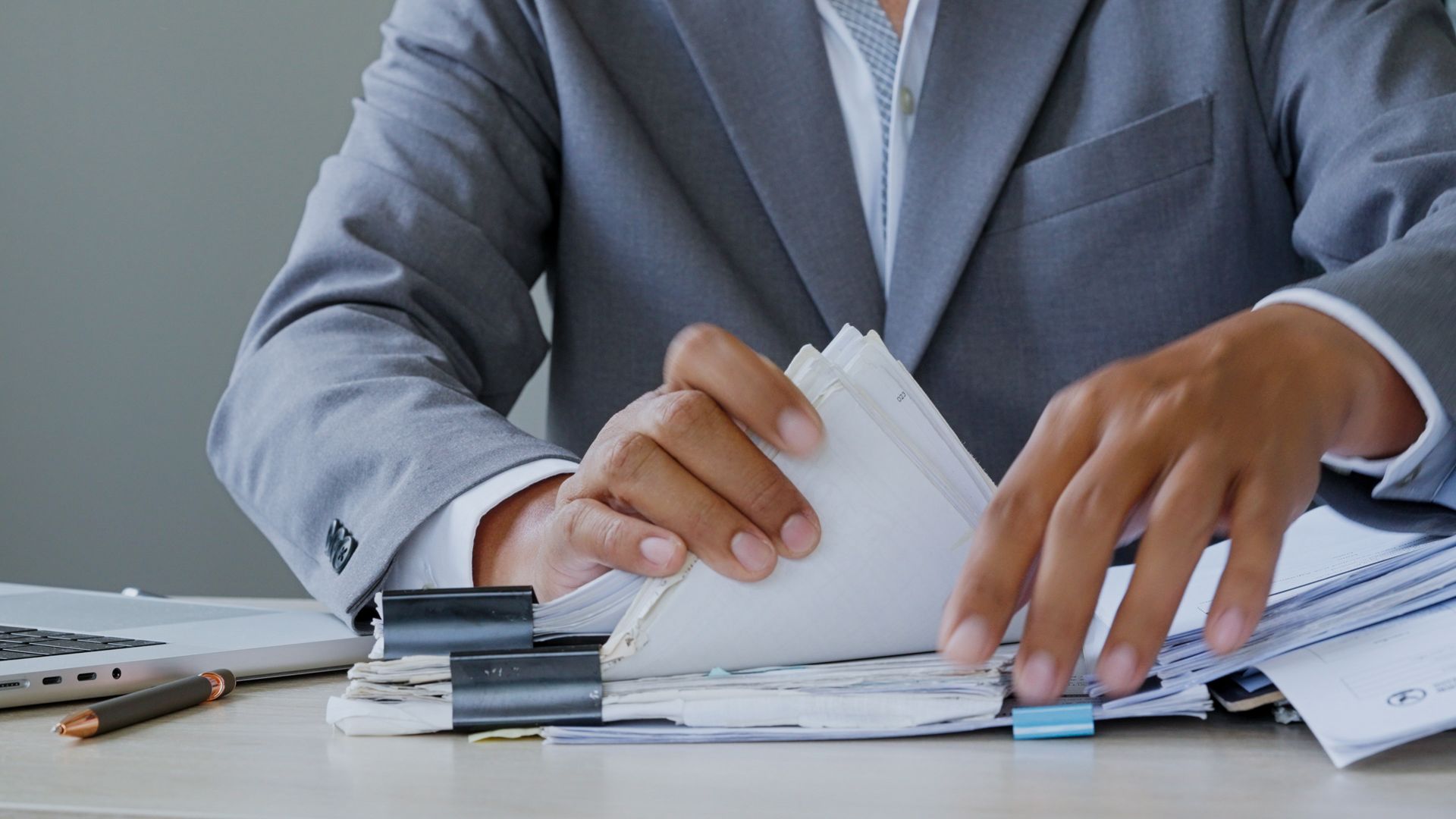 A businessman in a suit sorts through stacks of paperwork clipped at a desk.