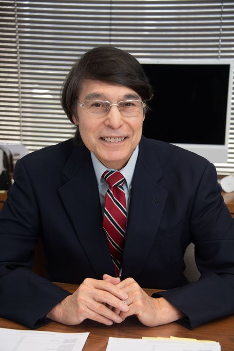 Man in suit at desk, smiling. Red and blue striped tie. Dark hair and glasses. Background includes blinds and a monitor.