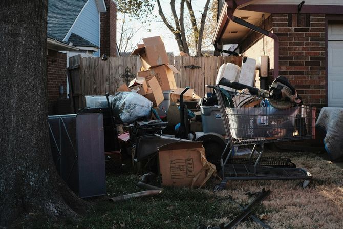Cluttered backyard with piled cardboard boxes, trash bags, and a shopping cart beside a house