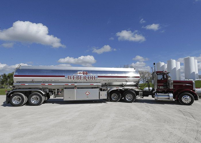 Maroon semi truck pulling a silver fuel tanker parked on a gravel lot under a blue sky