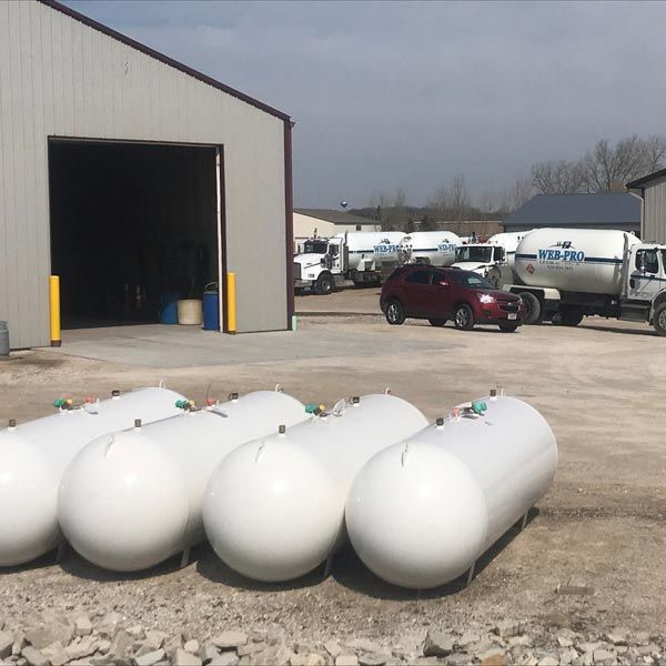 Four white propane tanks in a gravel yard beside a warehouse and fuel trucks
