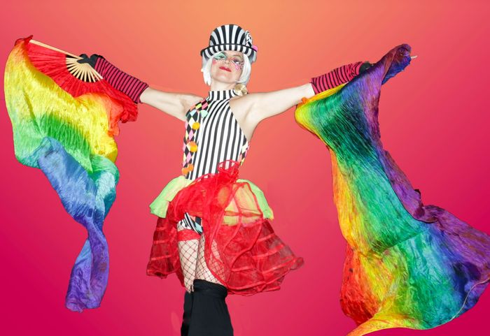 Person in clown-like costume with rainbow flags outstretched.