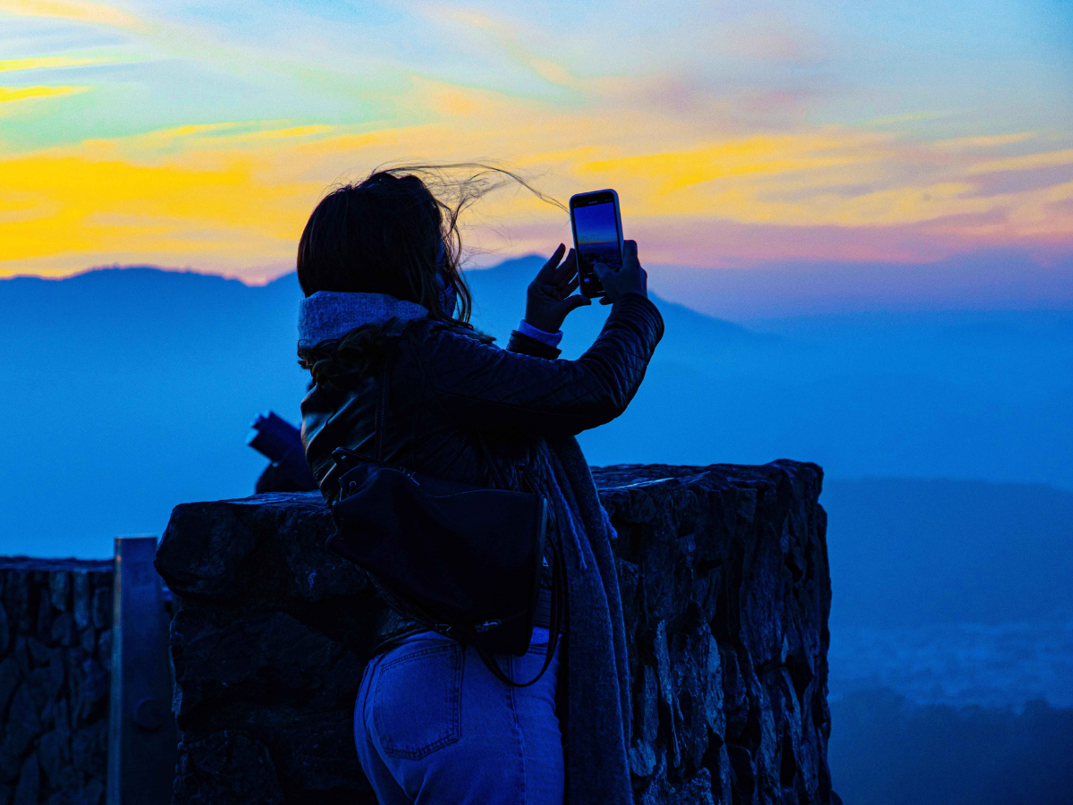 A silhouetted person standing by a stone wall holds up a phone to photograph a sunset over blue mountain ranges.