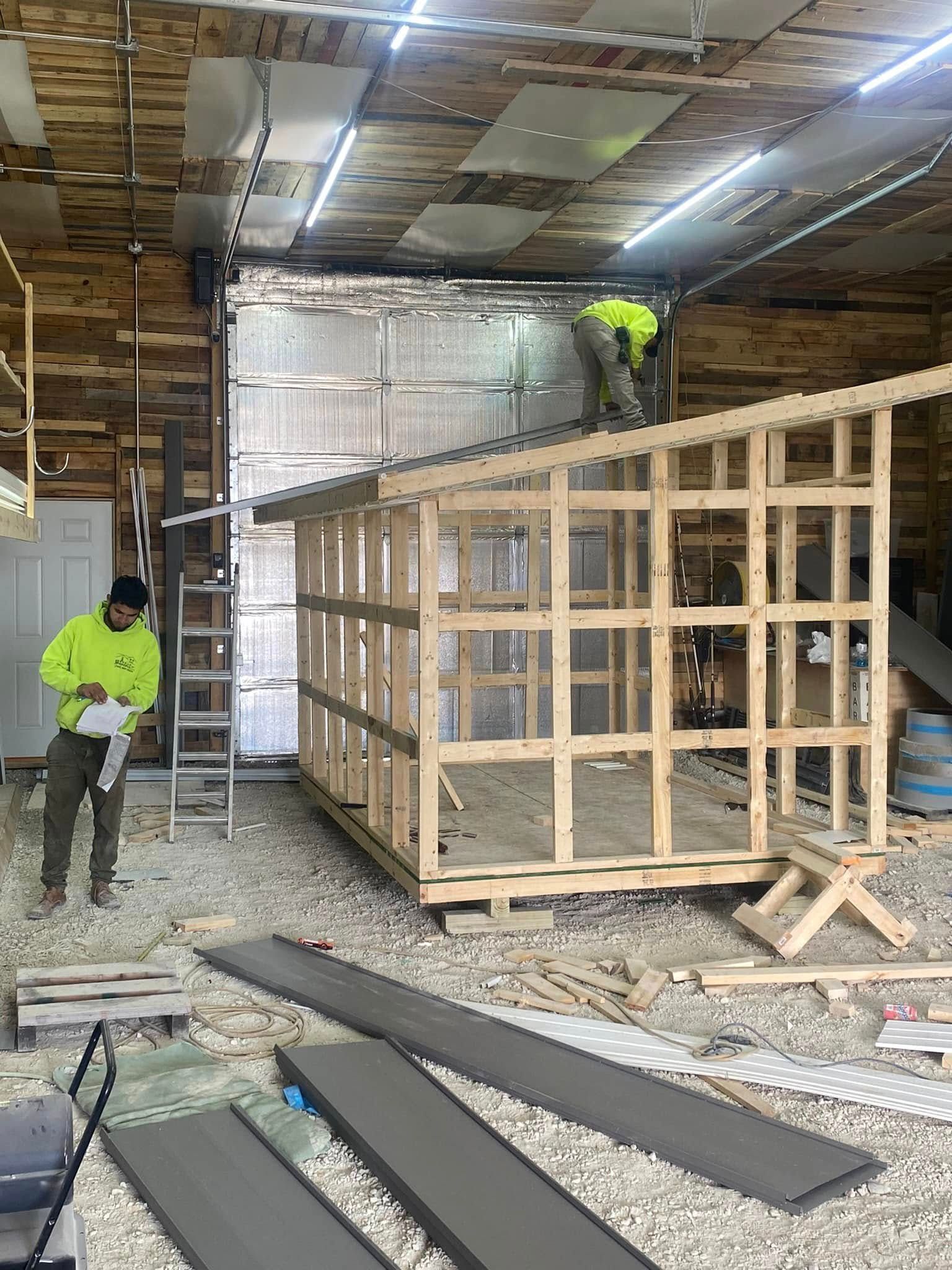 Workers building a wooden structure inside a warehouse with ladders and construction materials scattered nearby.