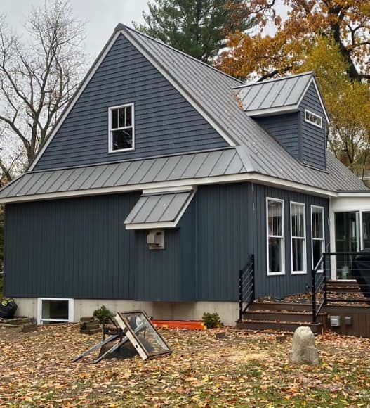 Blue-gray house with metal roof, dormer windows, and autumn leaves in the yard