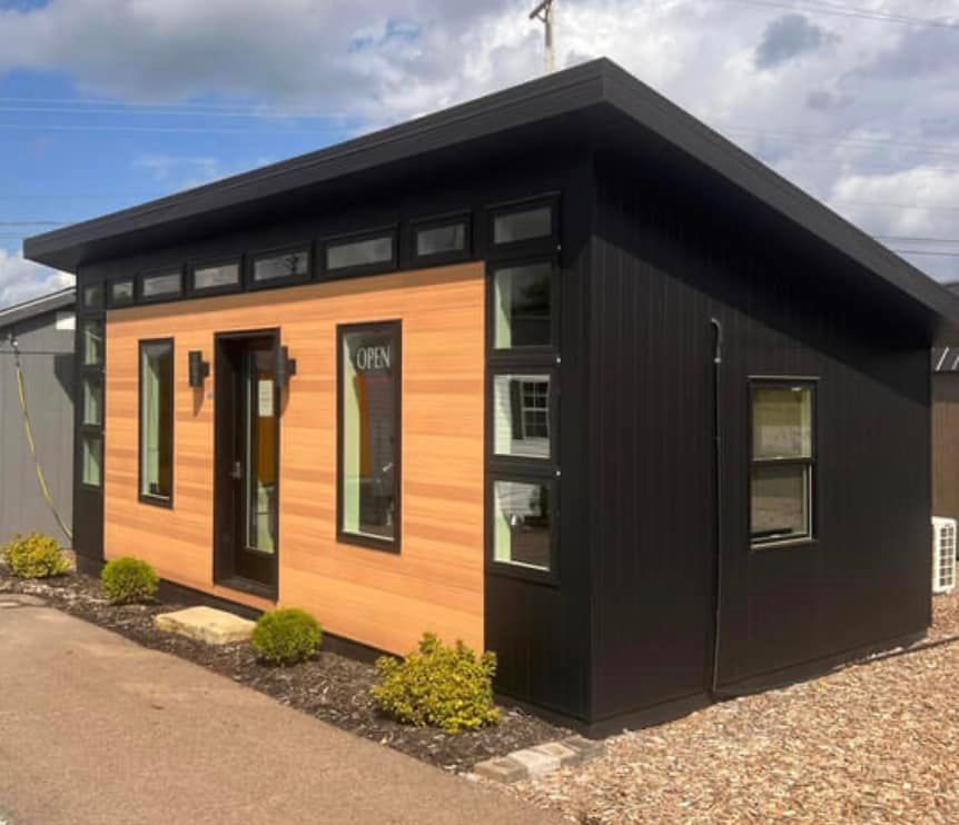 Small modern black-and-wood office building with large windows and a sloped roof under a cloudy sky