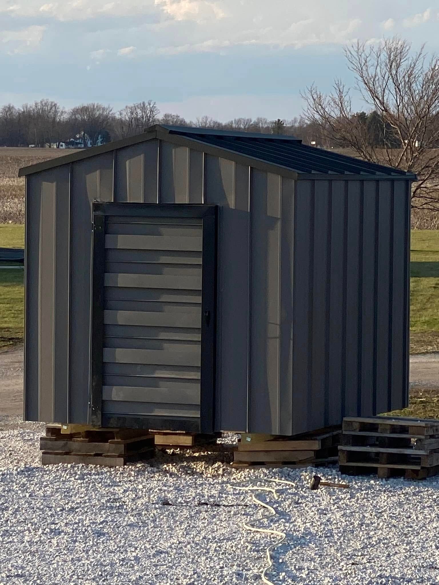 Small gray storage shed on pallets, with a closed corrugated door, sitting on gravel outdoors.